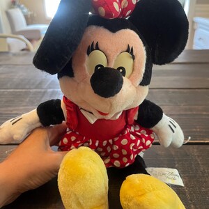 May include: A plush Minnie Mouse toy wearing a red dress with white polka dots and a red bow with white polka dots. The toy has yellow shoes and a "Walt Disney World" tag.