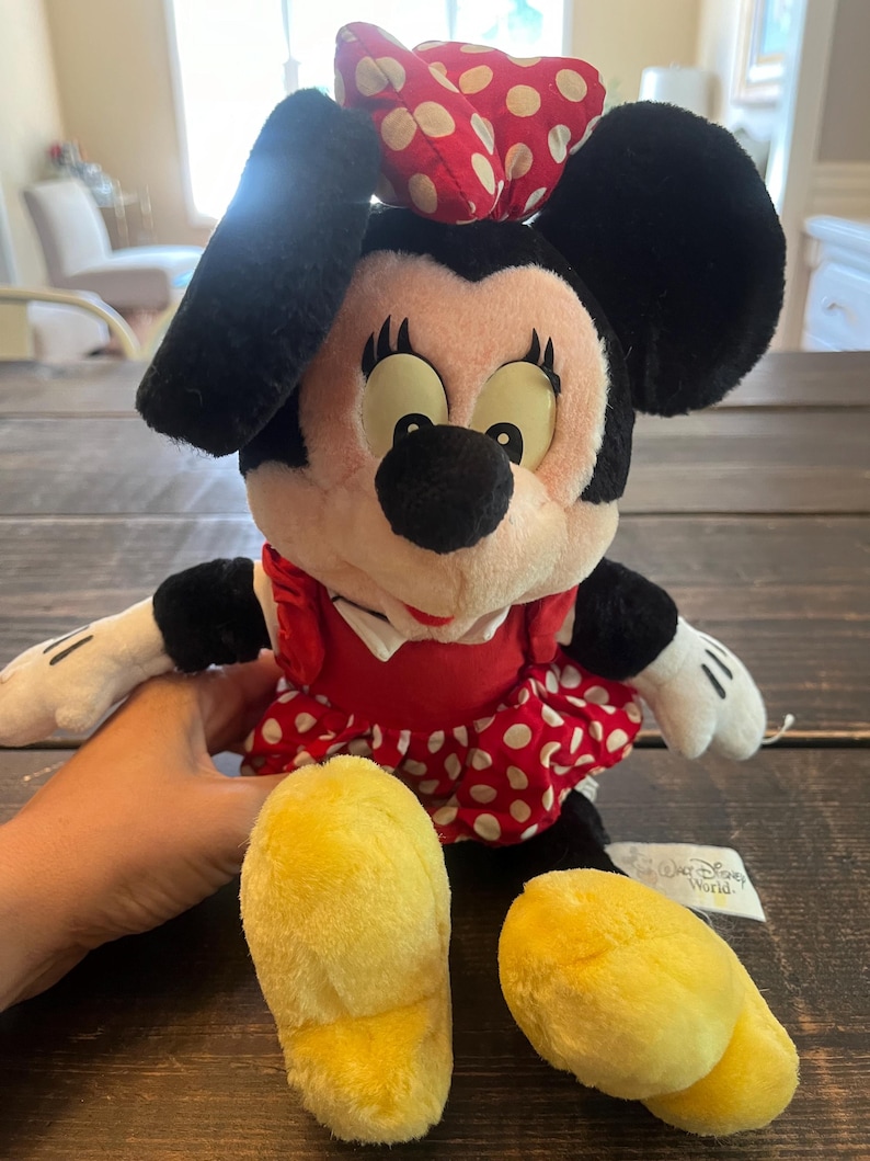 May include: A plush Minnie Mouse toy wearing a red dress with white polka dots and a red bow with white polka dots. The toy has yellow shoes and a "Walt Disney World" tag.