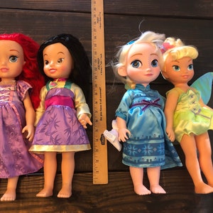 May include: Four Disney princess dolls, including Ariel, Mulan, Elsa, and Tinkerbell. The dolls are all wearing their signature dresses and have long hair.