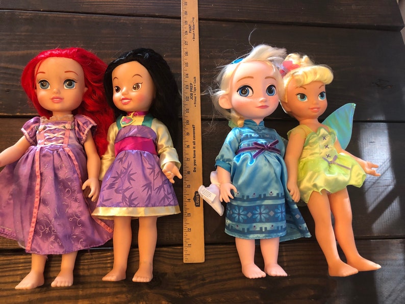 May include: Four Disney princess dolls, including Ariel, Mulan, Elsa, and Tinkerbell. The dolls are all wearing their signature dresses and have long hair.