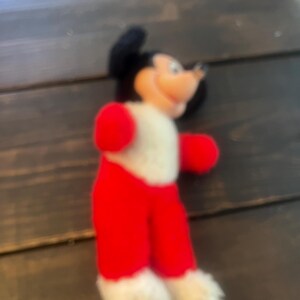 May include: A vintage Mickey Mouse plush toy. The plush features a black head with large ears, a white body, red shorts, red gloves, and white fluffy feet. The toy has a smiling expression and is made of soft fabric.
