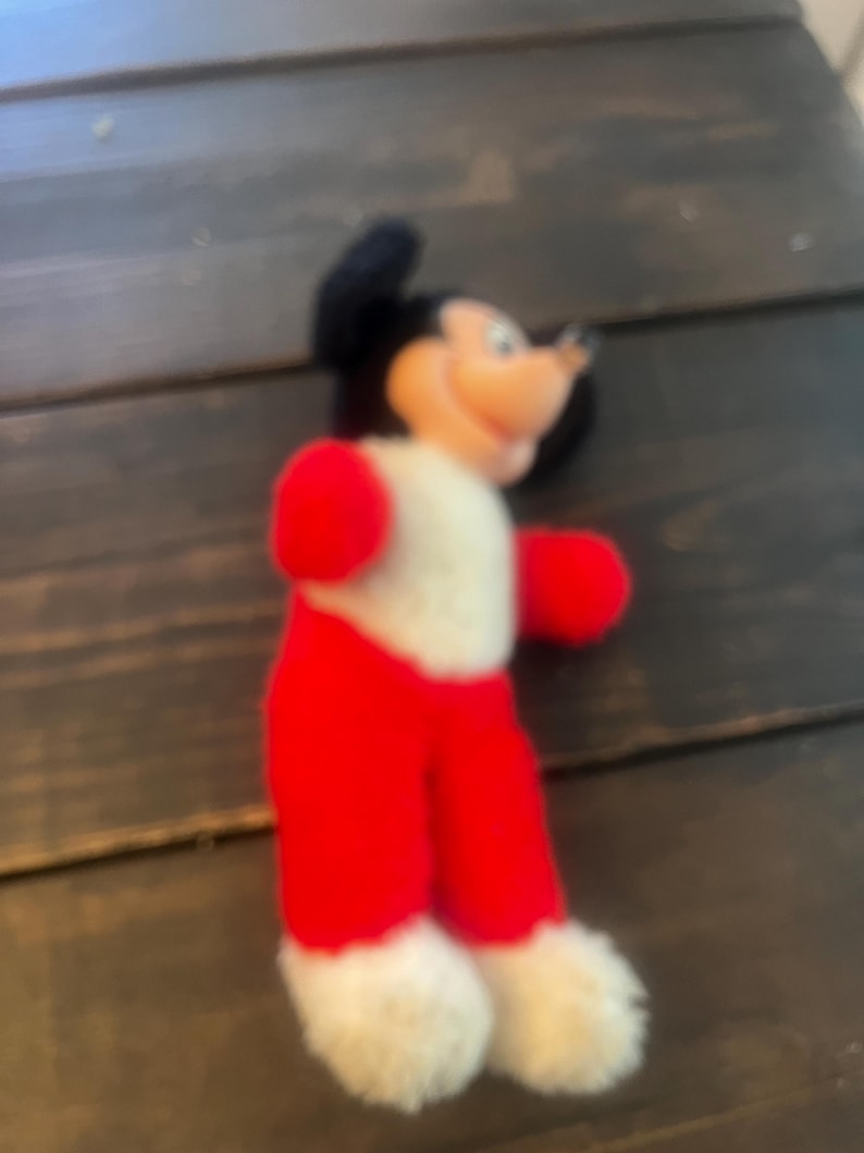 May include: A vintage Mickey Mouse plush toy. The plush features a black head with large ears, a white body, red shorts, red gloves, and white fluffy feet. The toy has a smiling expression and is made of soft fabric.