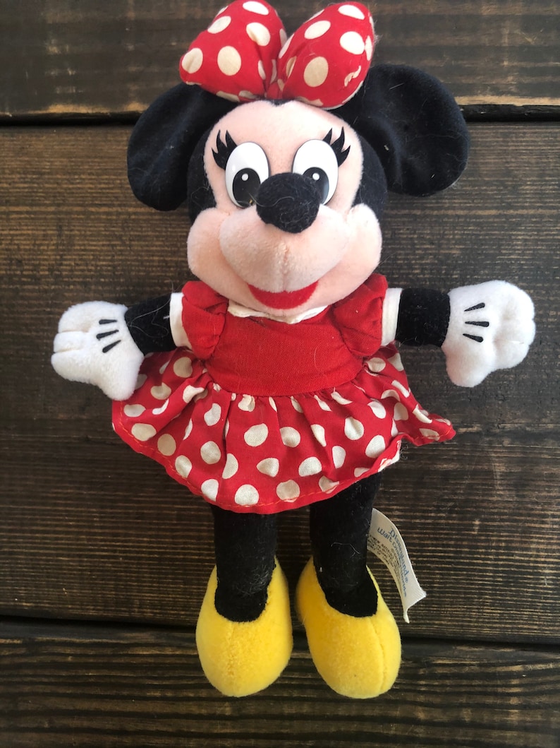 Puede incluir: Un vestido rojo con lunares blancos y un lazo rojo en la cabeza. El juguete de peluche es un personaje de dibujos animados con ojos negros y una nariz negra. El juguete lleva zapatos amarillos.