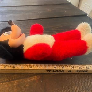 May include: A vintage plush toy of a cartoon character, featuring a black head, tan face, and red body with white accents. The toy is lying on a wooden surface. A wooden ruler is in the foreground.