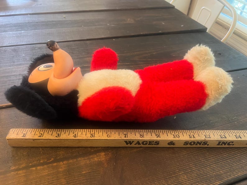 May include: A vintage plush toy of a cartoon character, featuring a black head, tan face, and red body with white accents. The toy is lying on a wooden surface. A wooden ruler is in the foreground.