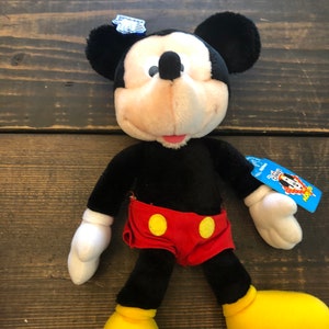 May include: Black and white plush Mickey Mouse toy wearing red shorts with yellow buttons. The toy has a blue tag with the words "Disney Plush" and a Mickey Mouse head logo.