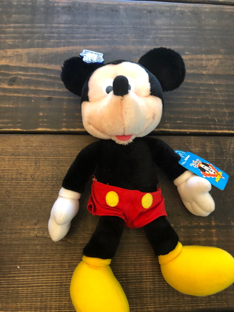 May include: Black and white plush Mickey Mouse toy wearing red shorts with yellow buttons. The toy has a blue tag with the words "Disney Plush" and a Mickey Mouse head logo.