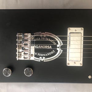 May include: A black cigar box guitar with silver hardware and strings. The guitar features a silver plate with the text "Fabrica de Tabacos Aganorsa A. Fernandez". Two black knobs are visible on the guitar's body.