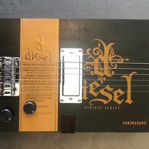 May include: A handmade cigar box guitar with a black body and a light brown neck. The guitar features the word "Diesel" in gold lettering, along with other text and a silver bridge and pickup.