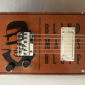 May include: A handmade cigar box guitar with a brown wooden body. The guitar features a black and white graphic of a man in a hat, the text "BLIND MANS BLUFF", chrome hardware, and black knobs. The neck is light-colored wood.