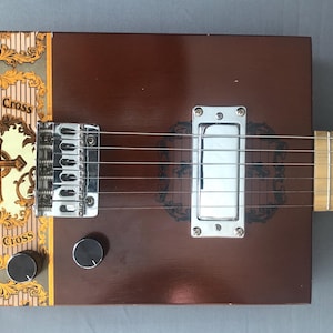 May include: A handmade cigar box guitar with a brown wooden body. The guitar features a silver bridge, pickup, and tuning knobs. The headstock is light-colored wood. The cigar box has the words "Bronze Cross" and a cross design.