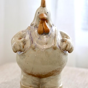 May include: A ceramic figurine of a chicken wearing overalls. The chicken has a glossy off-white glaze with brown accents on the comb, wattle, feet, and the edges of the overalls. It stands in an upright position with arms resting on its hips.