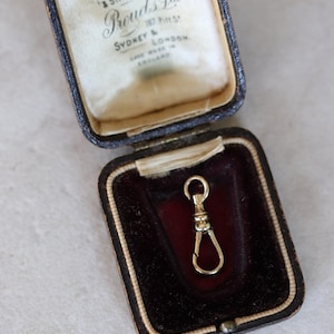 May include: A gold-toned metal swivel watch fob clasp resting inside a dark brown hinged jewellery box. The interior lid features printed text reading JEWELLERS & SILVERSMITHS, Prouds Ltd, 187 PITT ST, SYDNEY & LONDON, MADE IN ENGLAND.
