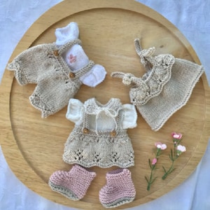 Puede incluir: Tres conjuntos de punto para una muñeca, que incluyen un mono, un vestido y una blusa sin mangas. Los conjuntos son beige y blanco con botones marrones. La muñeca no está en la imagen.