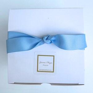 May include: A white square box tied with a light blue satin ribbon. A small sticker with gold trim and the text "Jeanne Player Fine Art" is affixed to the box. The website address jeanneplayerfineart.com is also visible.