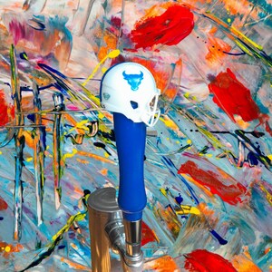 May include: A blue and white football helmet with a bull logo sits on top of a chrome beer tap. The background is a colorful abstract painting.