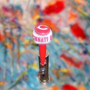 May include: A red and white baseball-shaped beer tap handle with the text "Cincinnati Reds" printed on it.