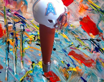 Puede incluir: Un tirador de cerveza marrón y blanco con forma de casco de fútbol americano con un logotipo azul y blanco de una torre de perforación de petróleo. El casco está sobre un fondo abstracto colorido.