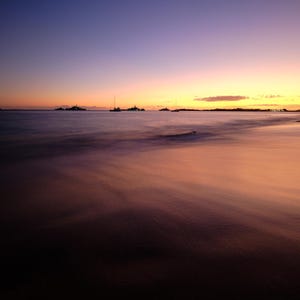 May include: A serene beach scene at dusk. The sky transitions from deep blue to vibrant orange and yellow hues. Silhouetted sailboats and distant landmasses are visible on the water. The foreground shows the wet sand reflecting the colorful sky.