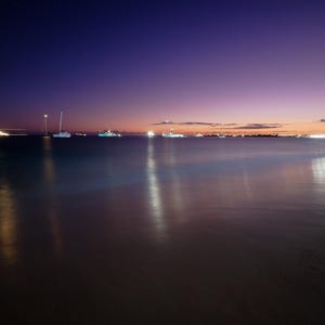 May include: A night time view of a calm body of water with a dark blue sky and a few stars.  The water reflects the lights of boats in the distance.