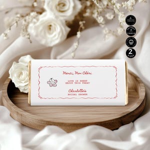 May include: A white chocolate bar with red text and a cherry illustration. The text reads "Merci, Mon Chéri", "Love is Sweet, Enjoy this Treat", and "Charlotte's Bridal Shower". The bar is on a wooden tray.