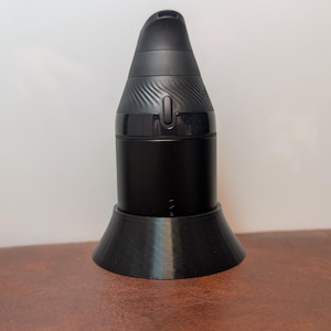 May include: A black, cylindrical device with a rounded top and a flared base. The top section has a textured design and a small button. The device is set on a matching black, conical stand. The overall design is sleek and modern.