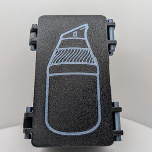 May include: A rectangular black storage case with a textured surface and two grey side latches. The front features a light blue outline graphic of a pump bottle with a nozzle and a ribbed collar, centered on the lid of the container.