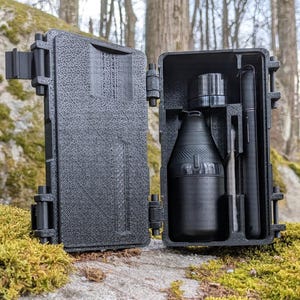 Puede incluir: Una caja negra abierta con un exterior texturizado, que revela una botella negra, un objeto cilíndrico y un objeto largo y delgado. La caja está sobre una roca cubierta de musgo en un entorno forestal.