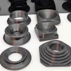 May include: A collection of various dark gray 3D-printed objects, including rings, cones, and geometric shapes. Some objects have a layered design, while others are solid. The objects are arranged on a white surface.