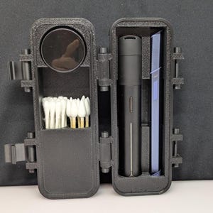 May include: An open, black, rectangular case with compartments. One side holds a black circular object and cotton swabs. The other side contains a black cylindrical object and a blue rectangular object. The case has a hinged design with latches.