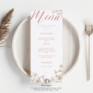 Może przedstawiać: Karta menu weselnego z kwiatowym wzorem, prezentowana na drewnianym talerzu z widelcem. Menu zawiera listę dań, w tym zupę dyniową, mostek wołowy i sznycel z kurczaka. Menu jest dla Fatimy i Yousufa, z datą 21.10.23.