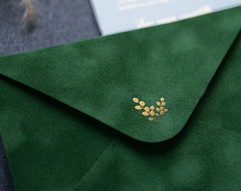 May include: A green velvet envelope with a gold foil design of leaves. The envelope is lying on a gray surface with a wax seal and a wax seal stamp.