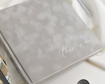 Puede incluir: Un libro de visitas cuadrado gris texturizado con las palabras "This is us" escritas en blanco. El libro está sobre una superficie blanca con una bandeja plateada y un sello de cera negro.