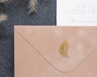 May include: A blush pink envelope with a gold leaf design. The envelope is lying on a dark gray surface with a white and brown wax seal and a wooden stamp.