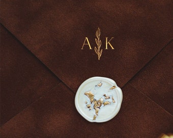 May include: A brown suede envelope with a gold wax seal and the initials "AK" in gold lettering. The envelope is on a white surface with white feathers and gold glitter.