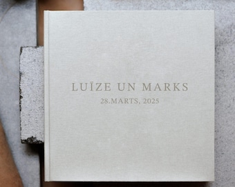 Puede incluir: Un álbum de fotos cuadrado con tapa dura recubierta de tela gris claro. La portada presenta el texto centrado y en relieve "LUĪZE UN MARKS" en una fuente serif, con la fecha "28.MARTS, 2025" impresa justo debajo en el mismo estilo.