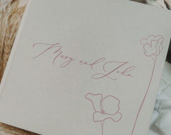 Puede incluir: Un álbum cuadrado cubierto de tela beige claro con el texto Mary and John en una delicada fuente cursiva de color rosa empolvado. Dos ilustraciones florales de líneas finas en el mismo color rosa se encuentran en el lado derecho.