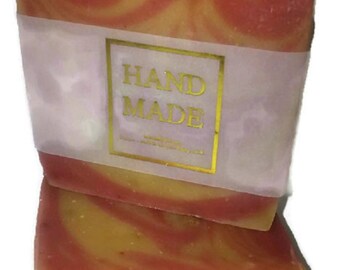May include: Two bars of handmade soap with a swirl pattern in shades of orange and red. The top bar has a white label with gold lettering that reads "HAND MADE".