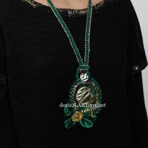 May include: A green and gold crocheted necklace with a large, ornate pendant featuring a silver mirror. The pendant is surrounded by green and gold crocheted flowers.