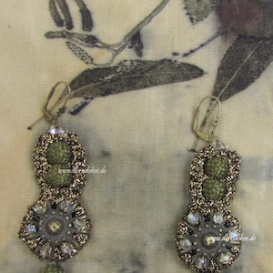 Peut inclure: Une paire de boucles d'oreilles vertes et dorées avec un motif floral. Les boucles d'oreilles sont en tissu crocheté et ont un centre perlé. Les boucles d'oreilles sont suspendues à un fond blanc.