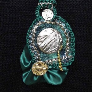 May include: A green crocheted pendant with a silver and green fabric circle in the center, surrounded by gold beads and a green satin bow. The pendant is attached to a green crocheted cord.