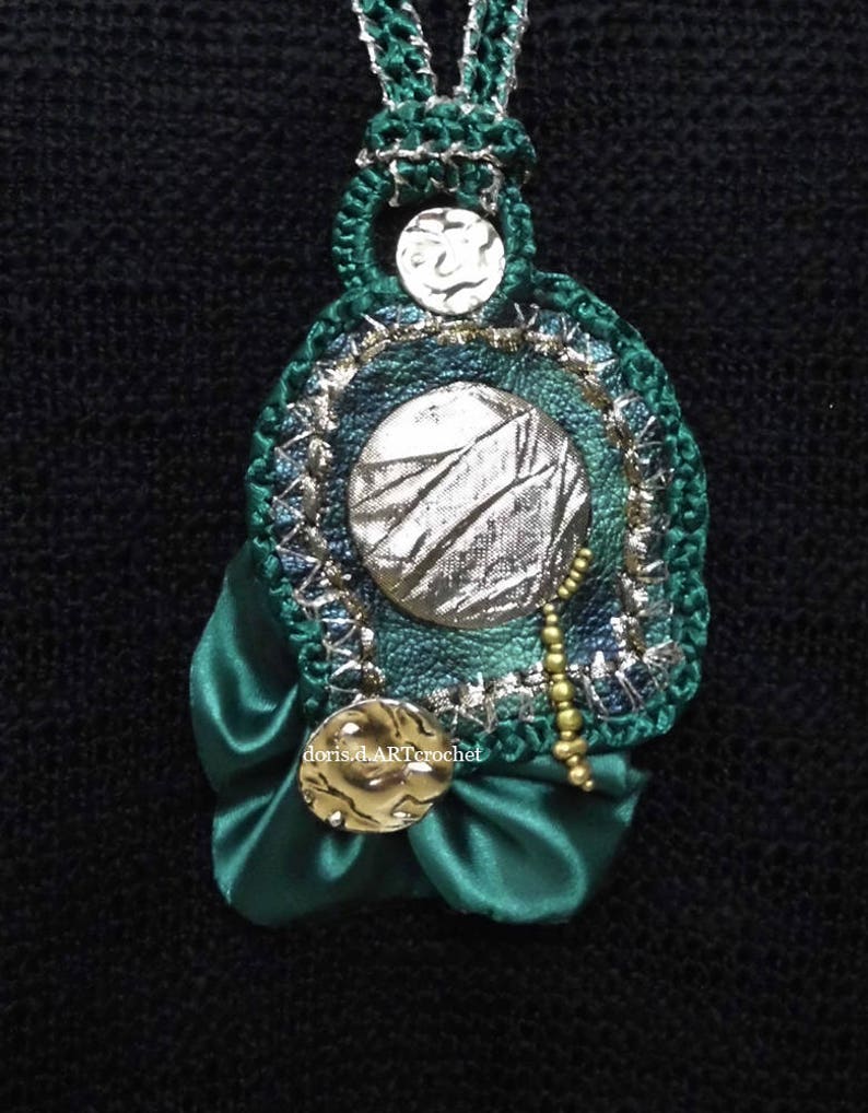 May include: A green crocheted pendant with a silver and green fabric circle in the center, surrounded by gold beads and a green satin bow. The pendant is attached to a green crocheted cord.