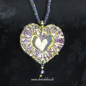May include: A heart-shaped pendant necklace with a gold and purple fabric design, featuring a smaller white heart in the center. The pendant is adorned with sequins and beads, and hangs from a dark blue cord.