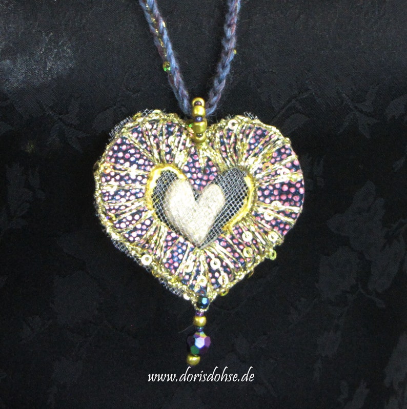 May include: A heart-shaped pendant necklace with a gold and purple fabric design, featuring a smaller white heart in the center. The pendant is adorned with sequins and beads, and hangs from a dark blue cord.