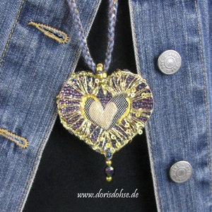 May include: A heart-shaped pendant necklace with a gold and purple beaded design. The heart is made of fabric and has a gold and white beaded design in the center. The necklace is hanging on a denim jacket.