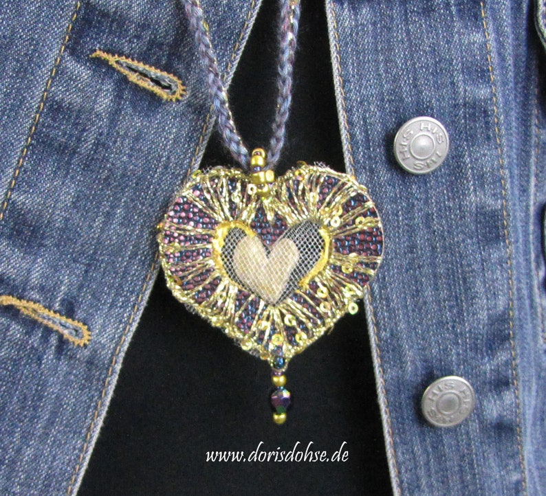 May include: A heart-shaped pendant necklace with a gold and purple beaded design. The heart is made of fabric and has a gold and white beaded design in the center. The necklace is hanging on a denim jacket.