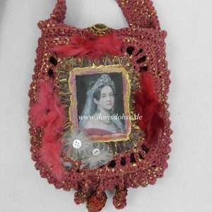 Könnte beinhalten: Eine kleine, gehäkelte Tasche in rosa und goldenen Farbtönen. Die Tasche zeigt ein gerahmtes Porträt einer Frau mit einer Krone und einem Schleier aus Spitze. Die Tasche hat auch rote Federn, Knöpfe und einen goldenen Quasten.