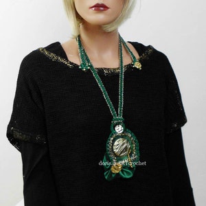 May include: A green and gold crocheted necklace with a large, round pendant. The pendant is made of crocheted circles and has a gold metallic center. The necklace is worn by a person with short blonde hair.