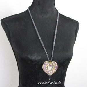 May include: A purple and gold heart-shaped pendant necklace with a beaded chain. The pendant has a delicate lace design and a small heart in the center. The necklace is displayed on a black mannequin.