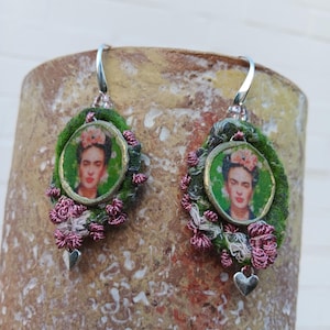Puede incluir: Pendientes hechos a mano con un retrato de Frida Kahlo. Presentan una base de tela verde, adornos florales rosas y pequeños dijes de corazón plateado. El retrato está enmarcado en dorado, creando un accesorio único.